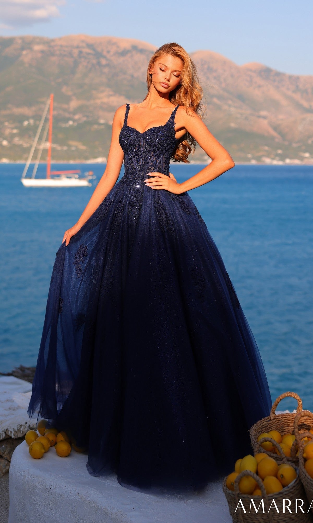 A woman in the Sheer-Corset Long Prom Dress: Amarra 89429 in navy stands by the seaside with mountains behind her and a basket of lemons nearby.